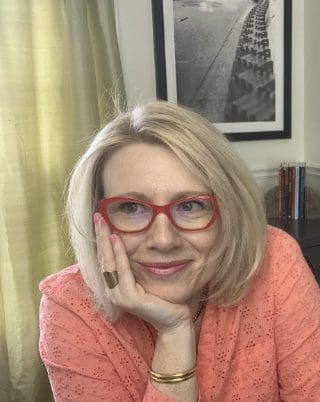 Dr Shannon Coates is sitting behind her desk and holding her head in her right hand. She is wearing red-rimmed reading glasses, a salmon-coloured, long-sleeved, collared shirt with the sleeves rolled up, two copper bangles, and a large brass ring on her ring finger. Dr C is smiling and looking off-camera to the right.