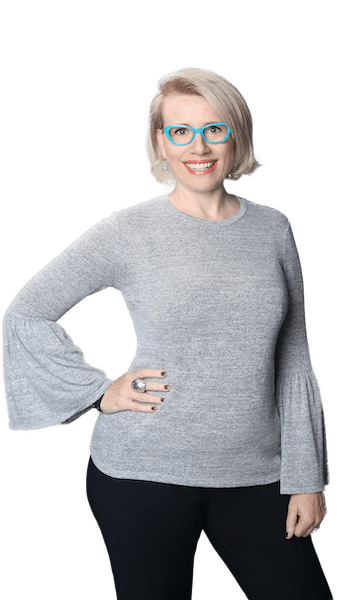 The Vocal Instrument 101 Follow Up Teacher Training Practicum creator, Dr. Shannon Coates, wearing teal-framed glasses, a grey shirt with wing arms, and black pants, strikes a jaunty pose with her right hand on her hip. She smiles openly at the camera.