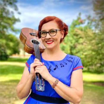 Testimonial photo of Ward, Marnie. She is wearing a blue dress and holding a ukulele over her shoulder.